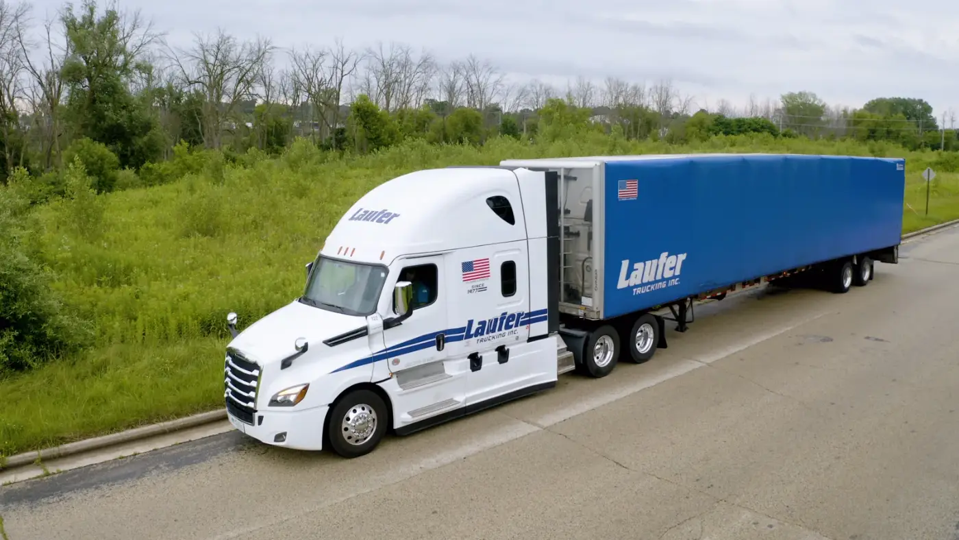 A white Laufer Trucking semi-truck pulls a blue curtain-side trailer on a highway to deliver freight.