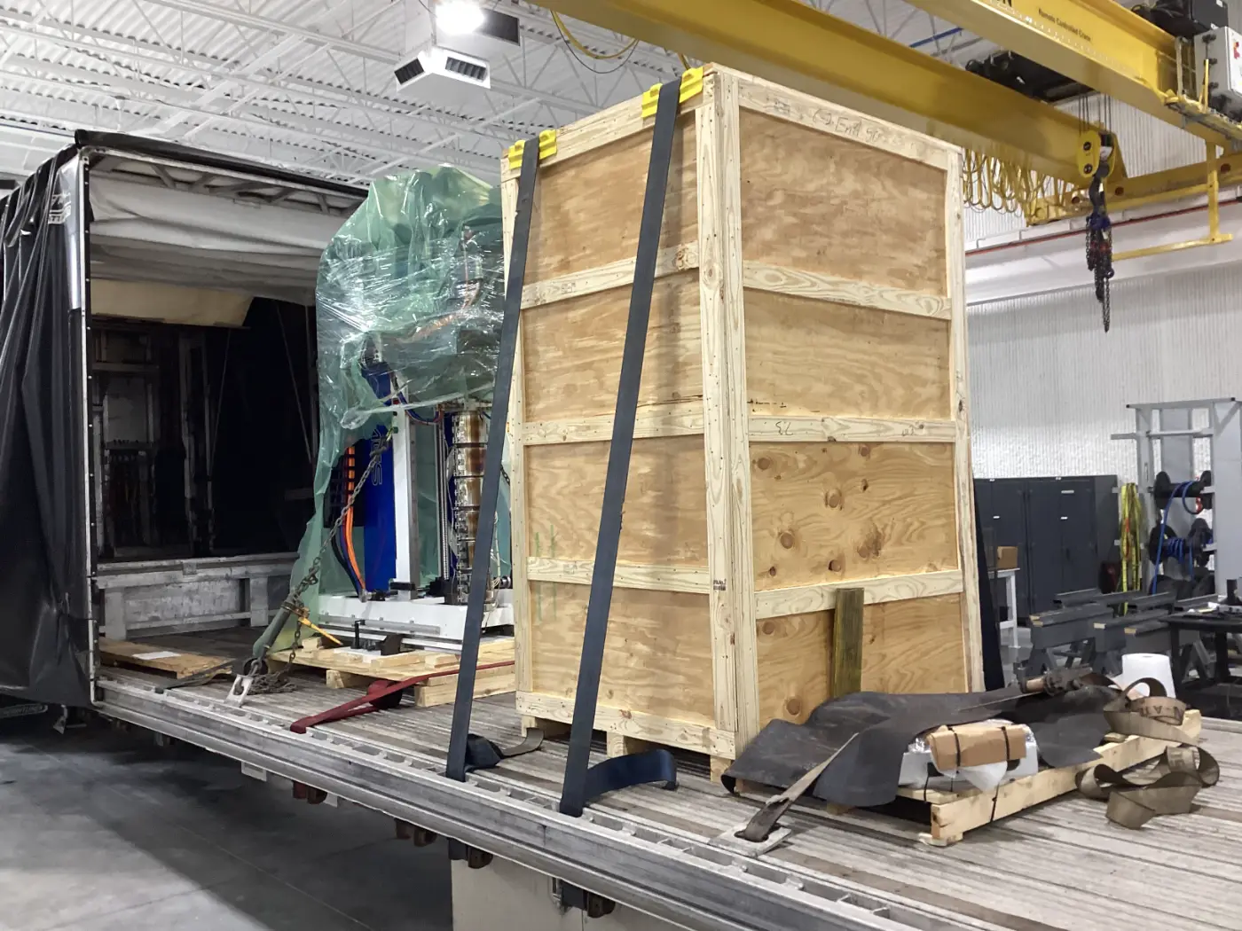 Tall heavy machinery and a large wooden crate secured with straps on a step deck Conestoga trailer in a warehouse.