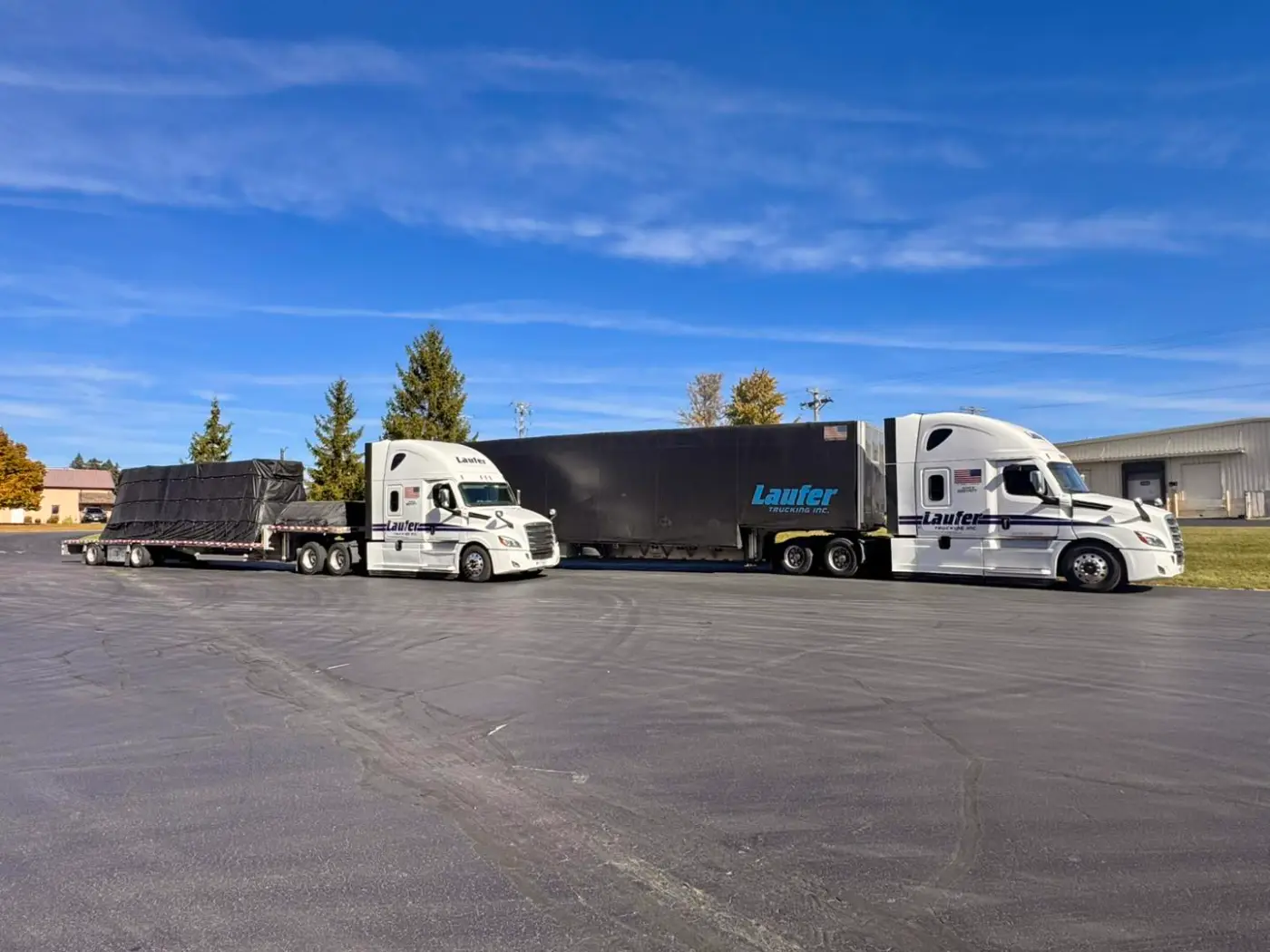 Two Laufer Trucking semi-trucks parked side by side, showcasing a flatbed trailer with a tarped load and a black curtain-side trailer.