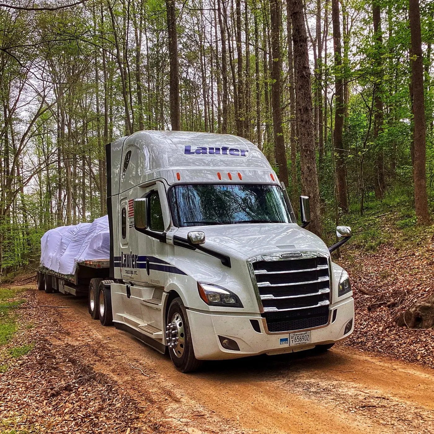 Laufer Trucking step deck trailer hauling tall machinery covered in white protective wrap through a wooded dirt road.