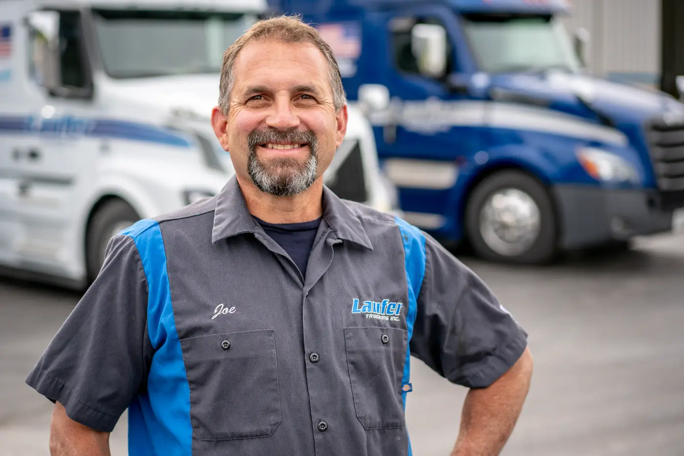 Smiling driver trainer Joe Volz stands in a Laufer Trucking uniform in front of white and blue semi trucks.