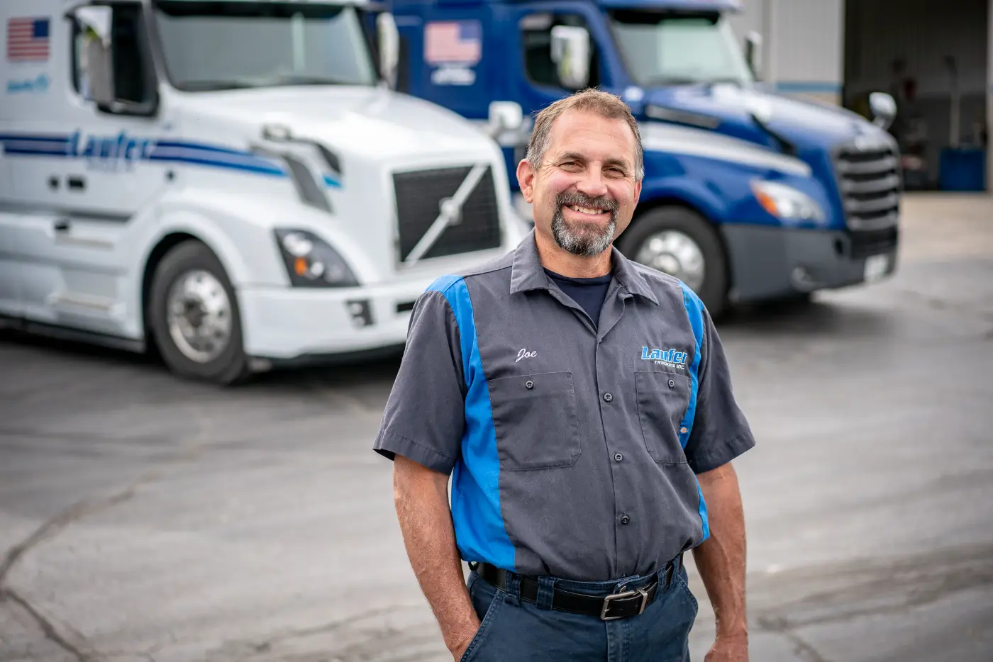 A smiling Laufer Trucking CDL driver standing in front of semicab trucks at a terminal in Wisconsin.