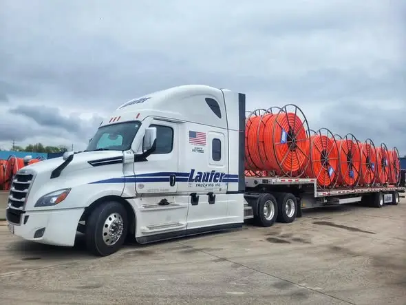 Laufer Trucking white semi-truck hauling large reels of orange industrial cable on a step deck trailer.
