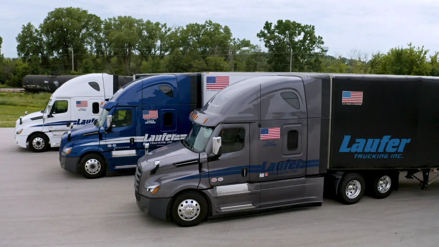 Three Laufer Trucking semi-trucks parked in a row, including a dry van and a Conestoga trailer with blue and grey cabs.