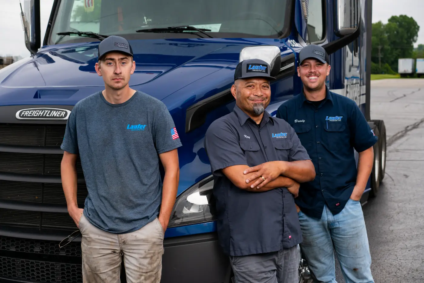 Three Laufer Trucking drivers standing in front of a blue Freightliner semi-truck, highlighting CDL career opportunities.