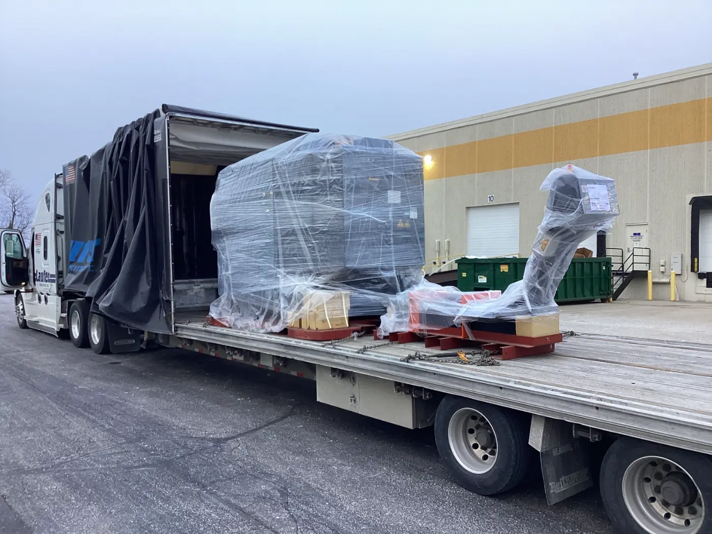 High-value industrial machinery secured for transport on a Laufer Trucking step deck Conestoga trailer with a retracted tarp system.