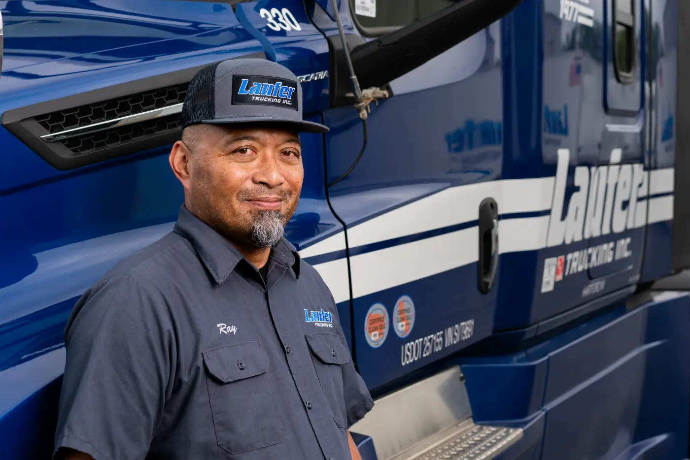 A smiling Laufer Trucking home-daily driver in uniform stands proudly in front of a blue semi-truck.