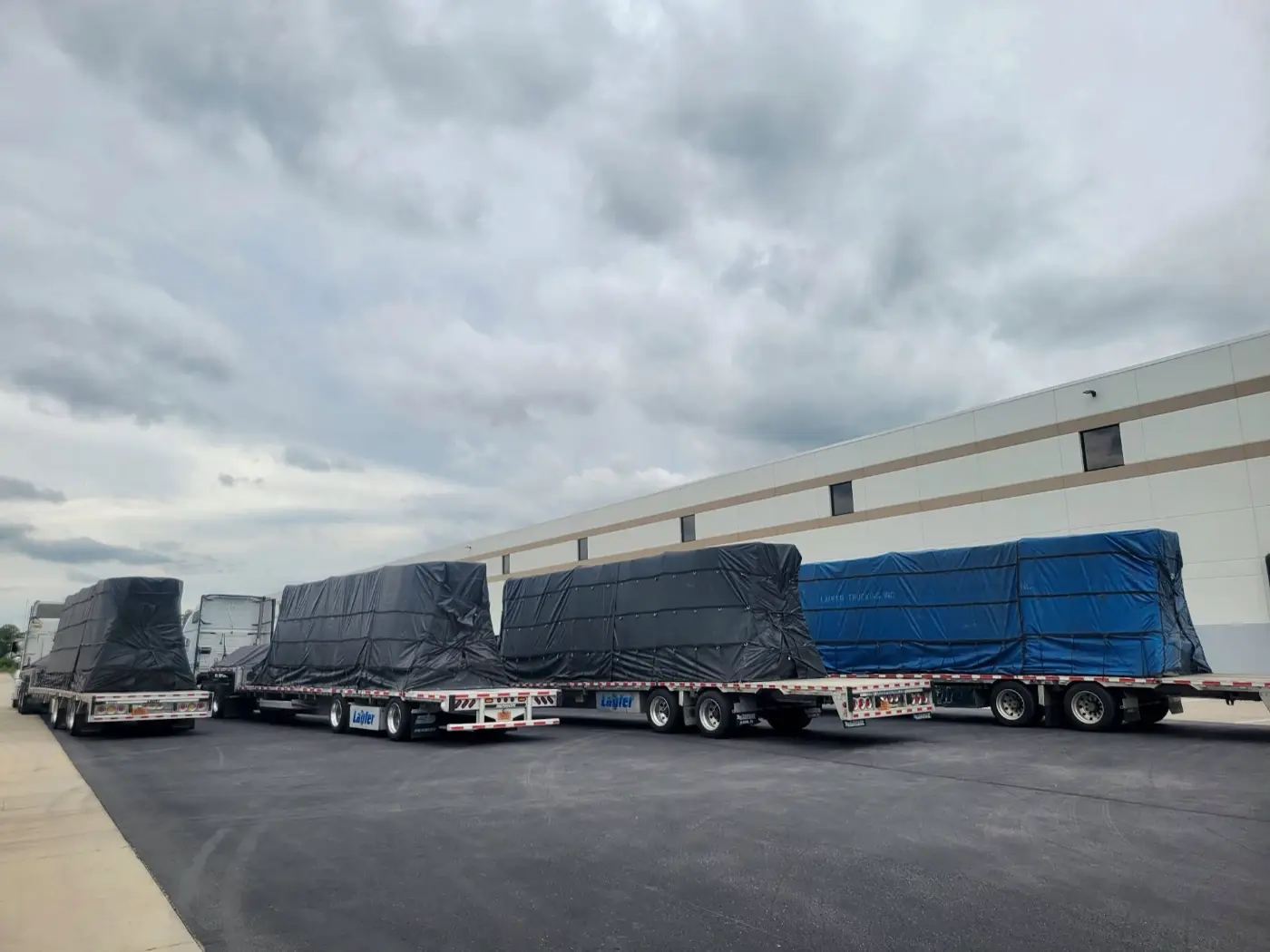 Three Laufer Trucking step deck trailers with large, tarped freight loads parked at a loading dock.