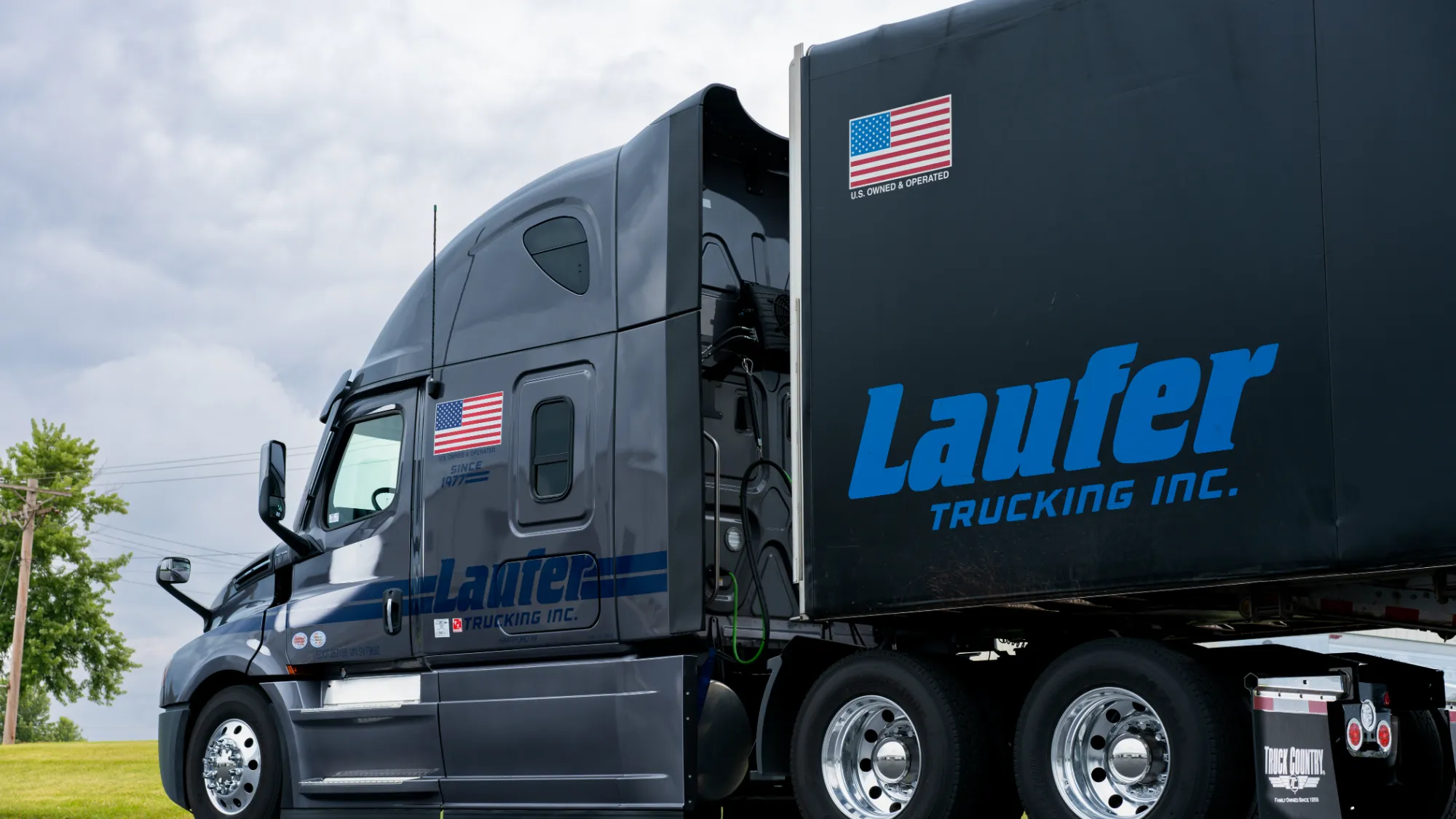 A charcoal gray Laufer Trucking semi-truck with a matching curtain-side trailer parked on a road with green trees in the background.