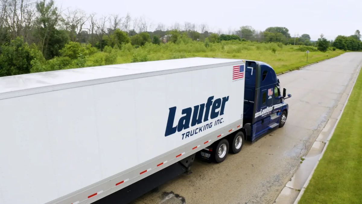 Blue Laufer Trucking semi truck hauling a white dry van trailer with special options logo on a paved road beside green trees.