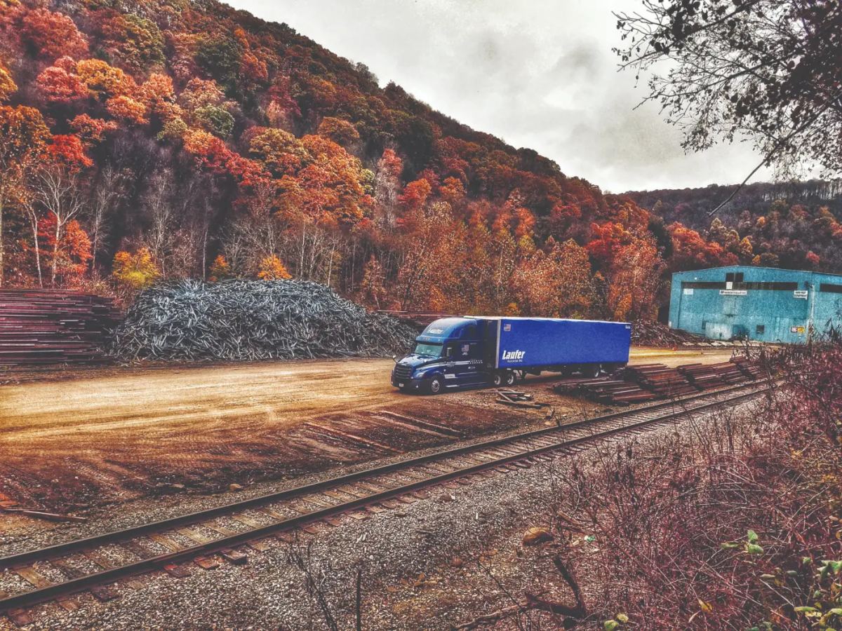 A Laufer Trucking curtain-side trailer parked at an industrial site for tooling and molding transport near an autumnal forest.