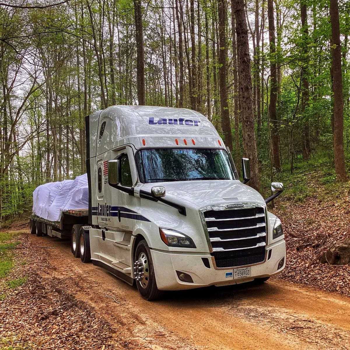 Laufer Trucking step deck trailer hauling tall machinery covered in white protective wrap through a wooded dirt road.