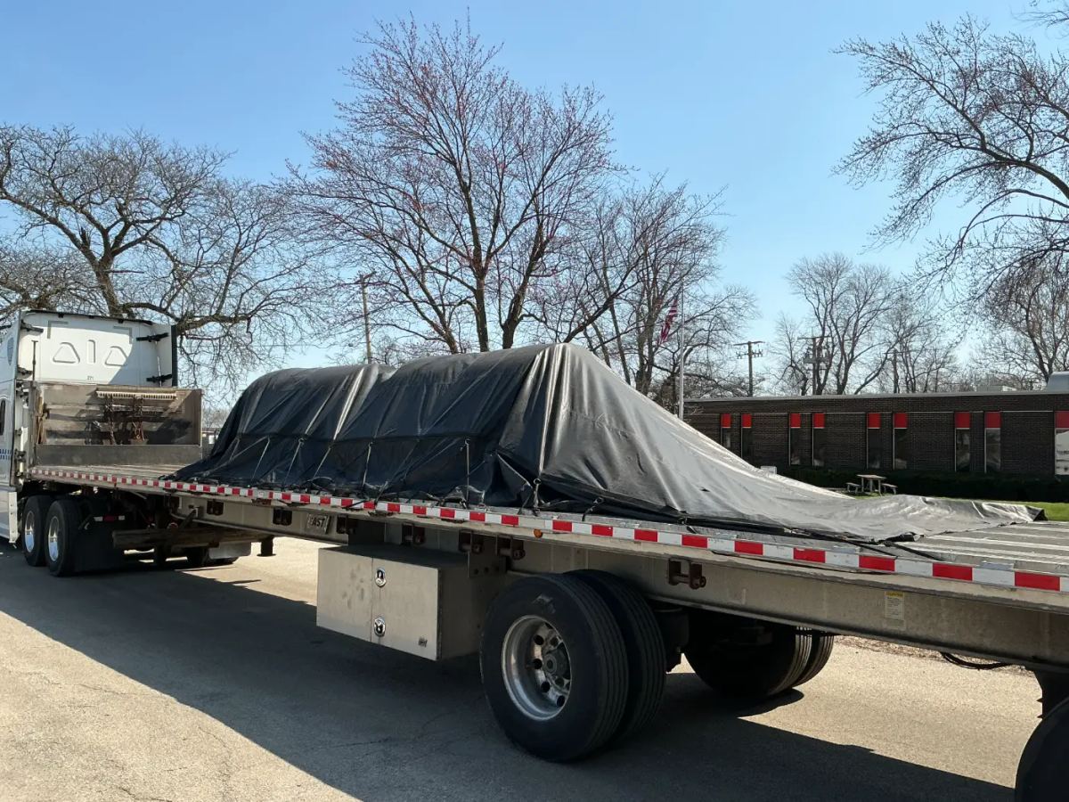A Laufer Trucking flatbed trailer transporting construction materials secured under a heavy-duty black weather-protective tarp.