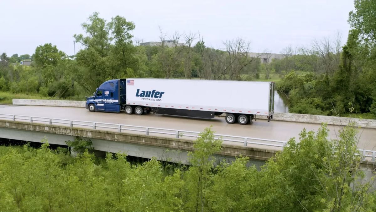 A Laufer Trucking blue semi-truck pulls a white dry van trailer over a bridge, providing transport for packaged food and beverage freight.