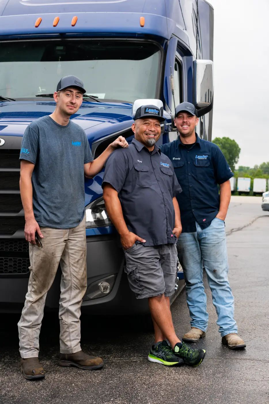Three smiling Laufer Trucking team members stand together in front of a blue semi-truck in the company yard.