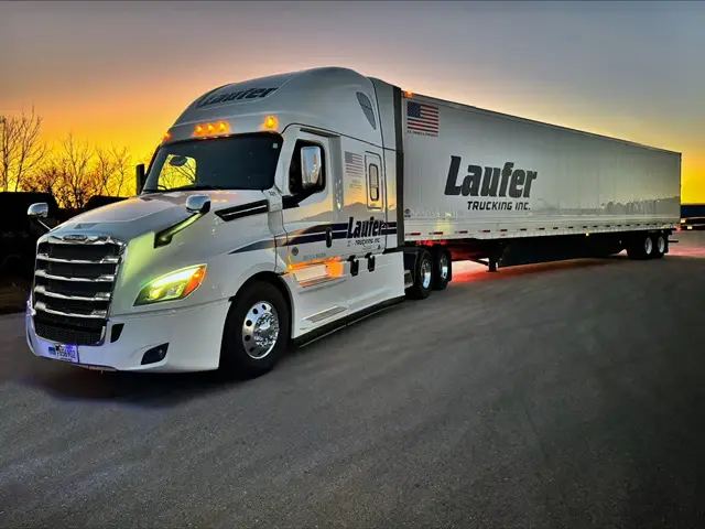 A white Laufer Trucking dry van semi-truck parked on an asphalt lot at sunset.
