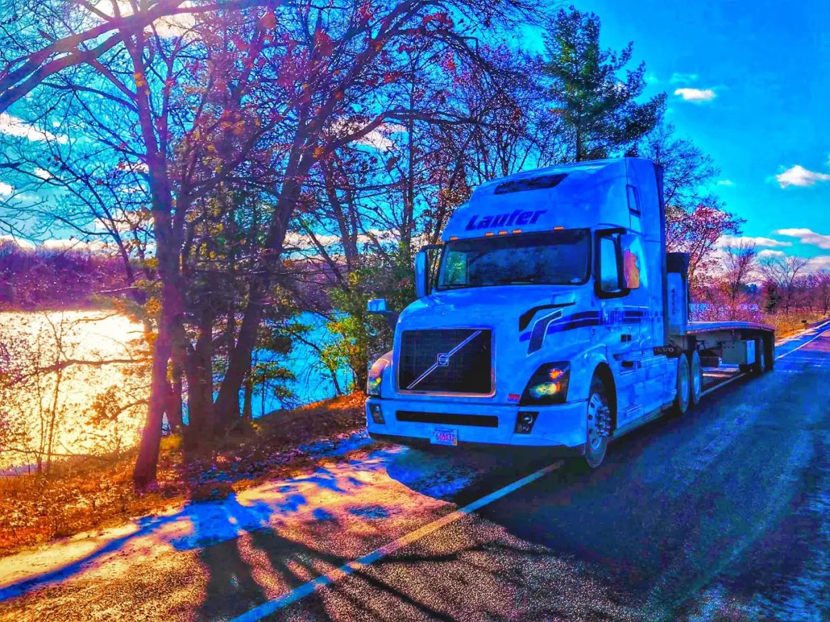 A white Laufer Trucking flatbed semi-truck parked on a scenic roadside near a lake, ideal for hauling raw steel and heavy freight.