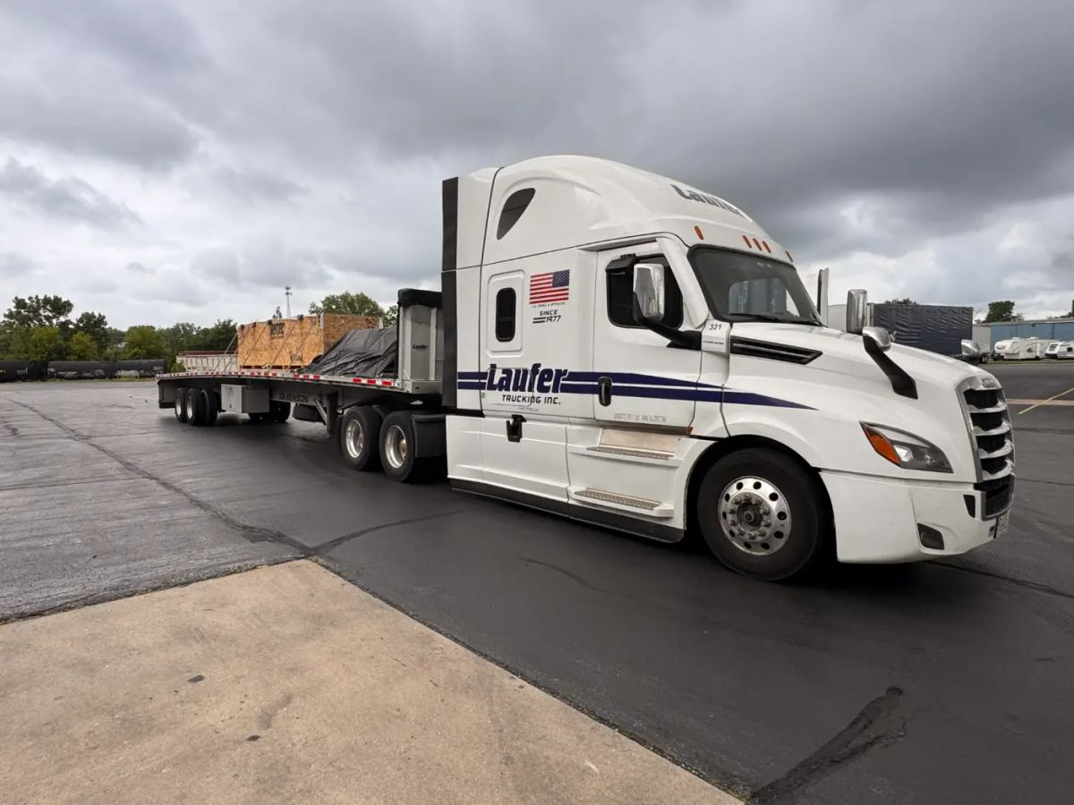 Laufer Trucking white semi-truck with an open flatbed trailer hauling heavy equipment and a large wooden crate.
