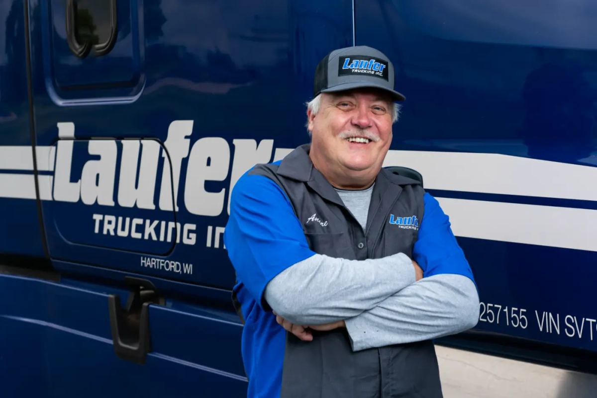 A smiling Laufer Trucking driver wearing a company hat and uniform stands with arms crossed in front of a blue semi-truck in Hartford.