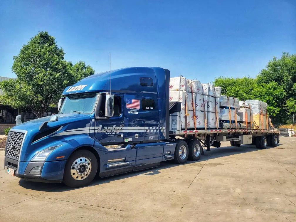 A blue Laufer Trucking semi-truck hauls long-haul freight on an open flat trailer secured with orange straps under a clear blue sky.
