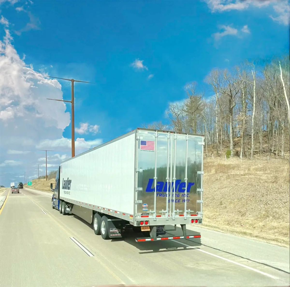 A blue Laufer Trucking semi-truck with a white dry van trailer transporting boxed consumer goods along a wooded highway.