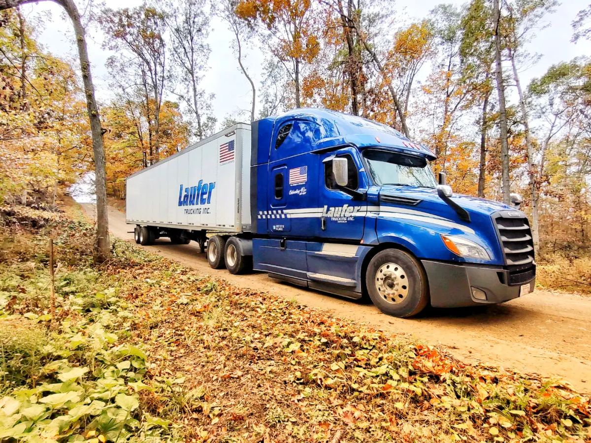 A blue Laufer Trucking semi-truck pulls a white dry van trailer along a narrow dirt road surrounded by autumn foliage.