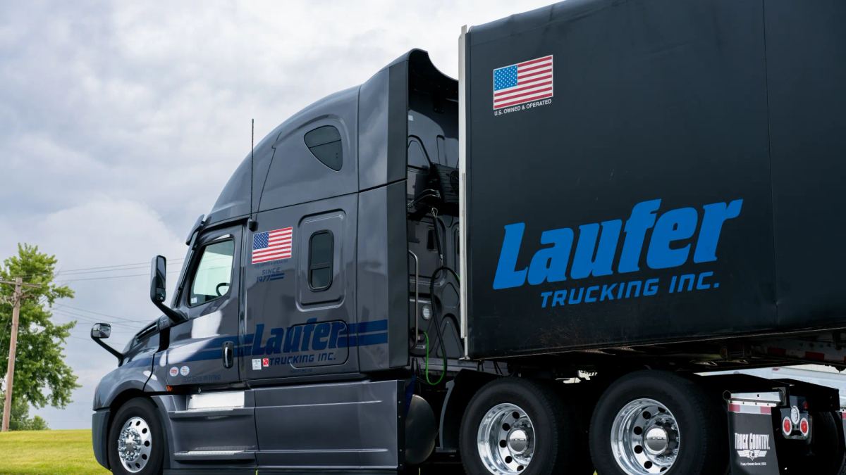 A charcoal gray Laufer Trucking semi-truck with a matching curtain-side trailer parked on a road with green trees in the background.