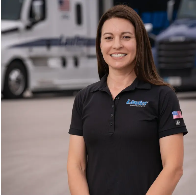 Krista Laufer smiling in a black Laufer Trucking polo shirt with a semi-truck blurred in the background.